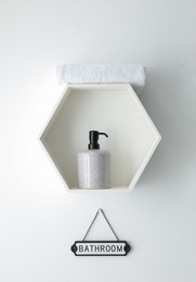 Photo of Stylish hexagon shaped shelf with towel and soap dispenser above Bathroom sign on white wall. Interior element