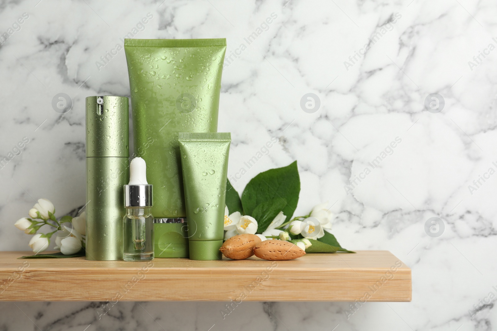 Photo of Set of cosmetic products, almond nuts and flowers on wooden shelf near white marble wall