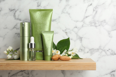 Set of cosmetic products, almond nuts and flowers on wooden shelf near white marble wall