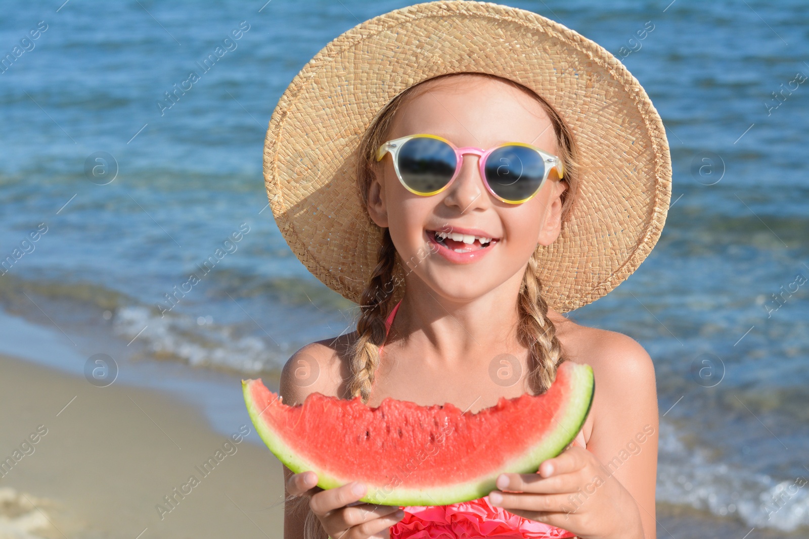 Photo of Cute little girl in straw hat and sunglasses eating juicy watermelon on beach