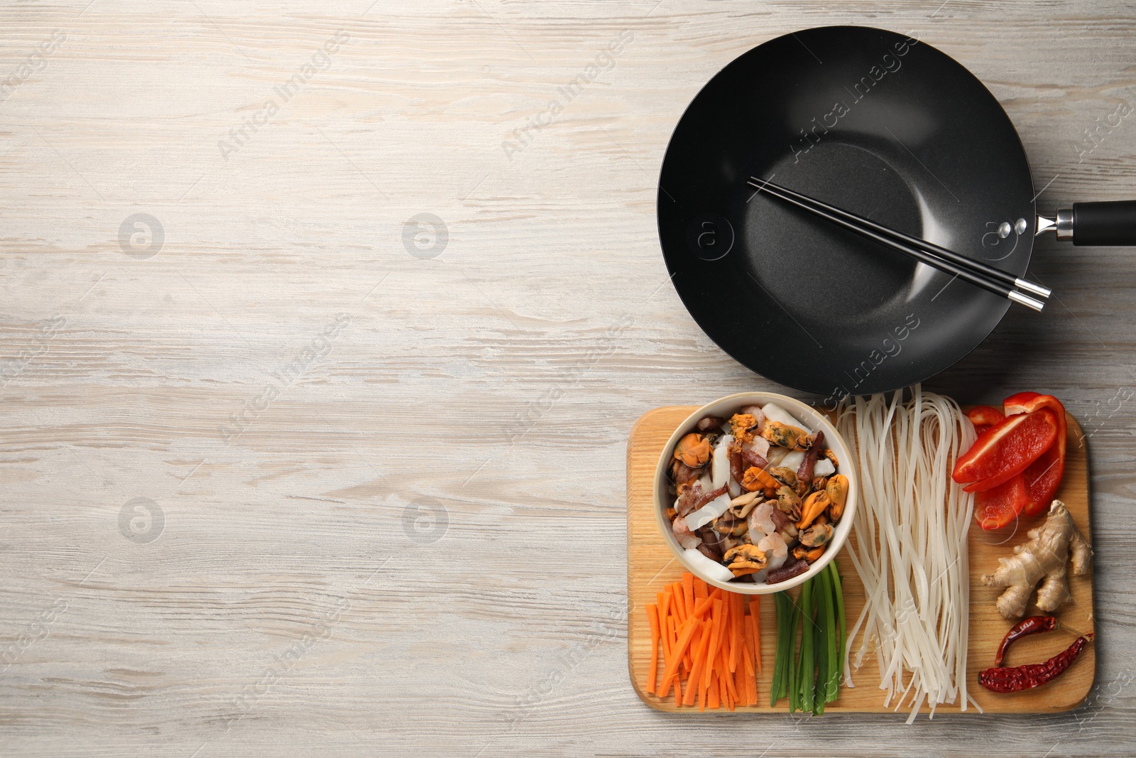 Photo of Black wok, chopsticks and board with products on light wooden table, top view. Space for text