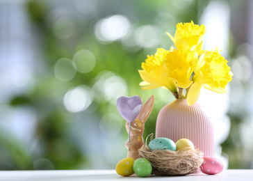 Photo of Painted Easter eggs, ceramic bunny and flowers on white table against blurred background. Space for text