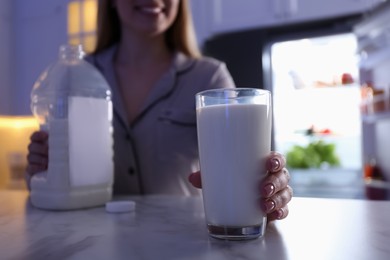Young woman holding glass and gallon bottle of milk on white marble table in kitchen at night, closeup