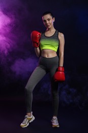 Photo of Beautiful woman wearing boxing gloves in color lights and smoke on black background