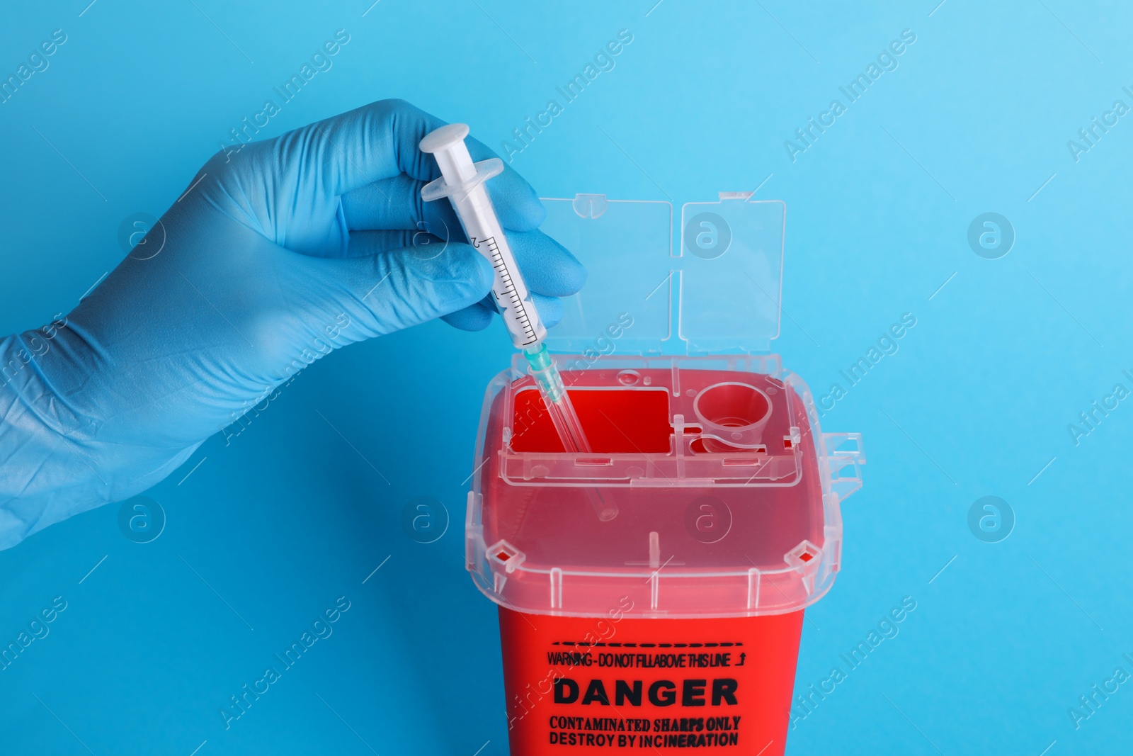 Photo of Doctor throwing used syringe into sharps container on light blue background, closeup