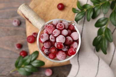 Photo of Frozen red cranberries in glass pot and green leaves on wooden table, top view