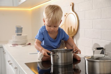 Photo of Curious little boy playing with pot on electric stove in kitchen