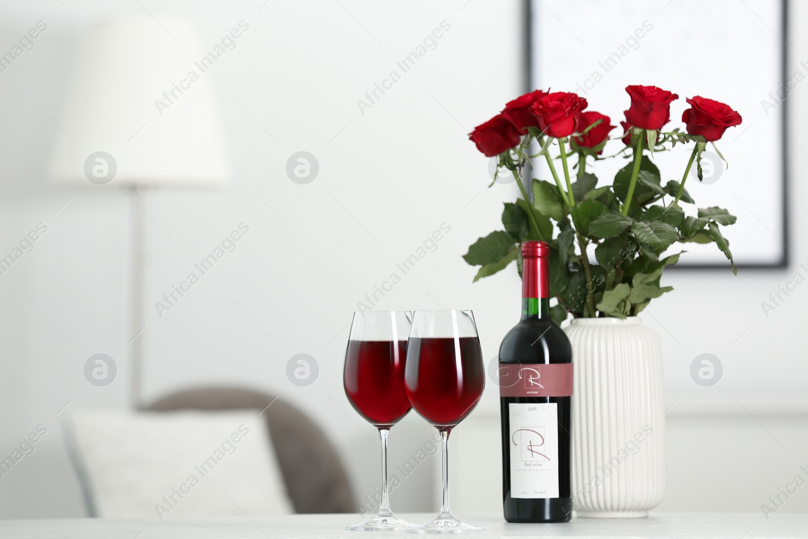 Photo of Bottle, glasses of red wine and vase with roses on table in room, space for text. Romantic date