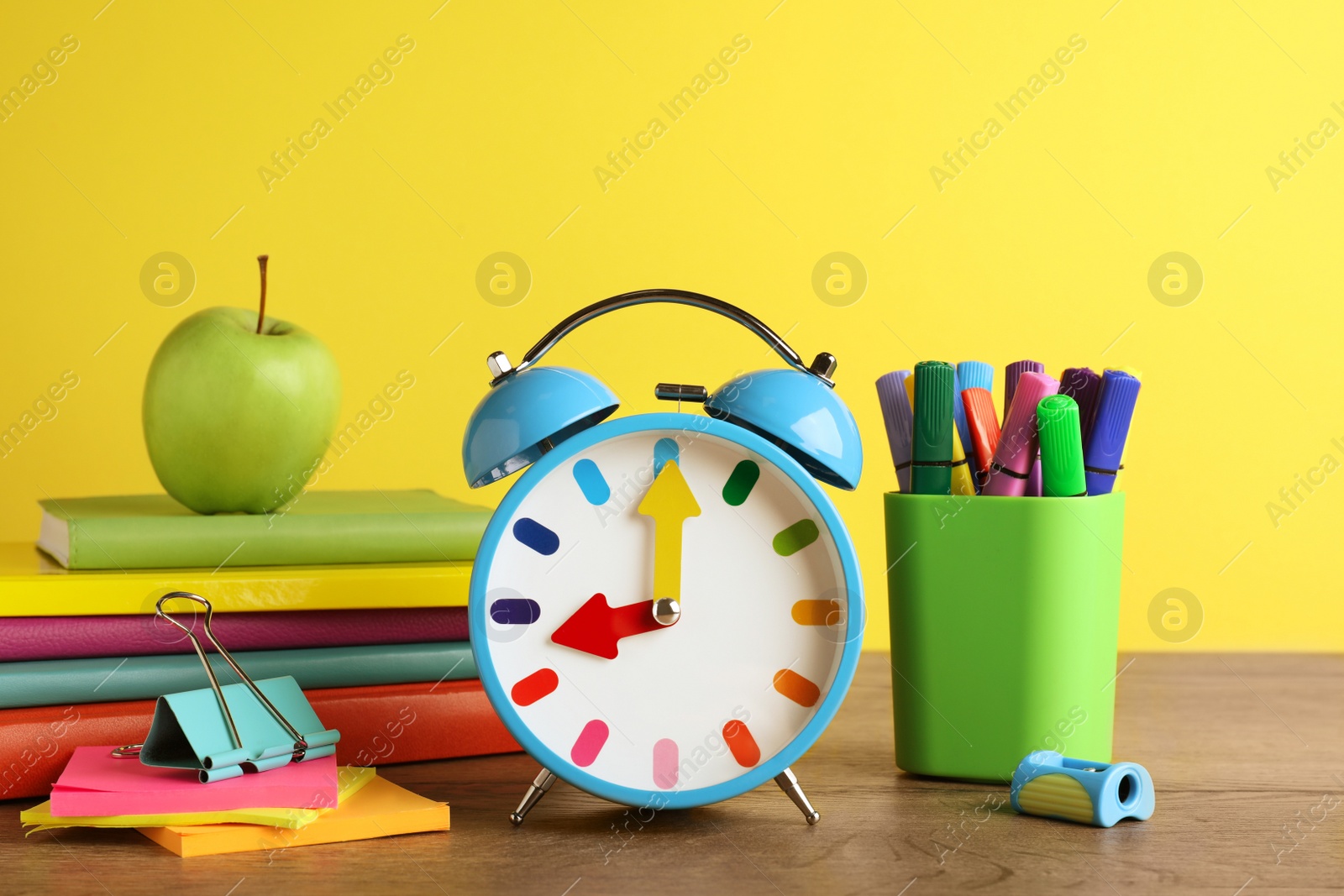 Photo of Light blue alarm clock and different stationery on wooden table against yellow background. School time