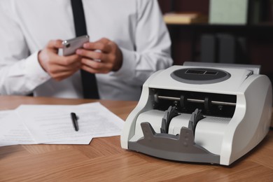 Photo of Modern banknote counter and blurred view of man using smartphone at wooden table indoors