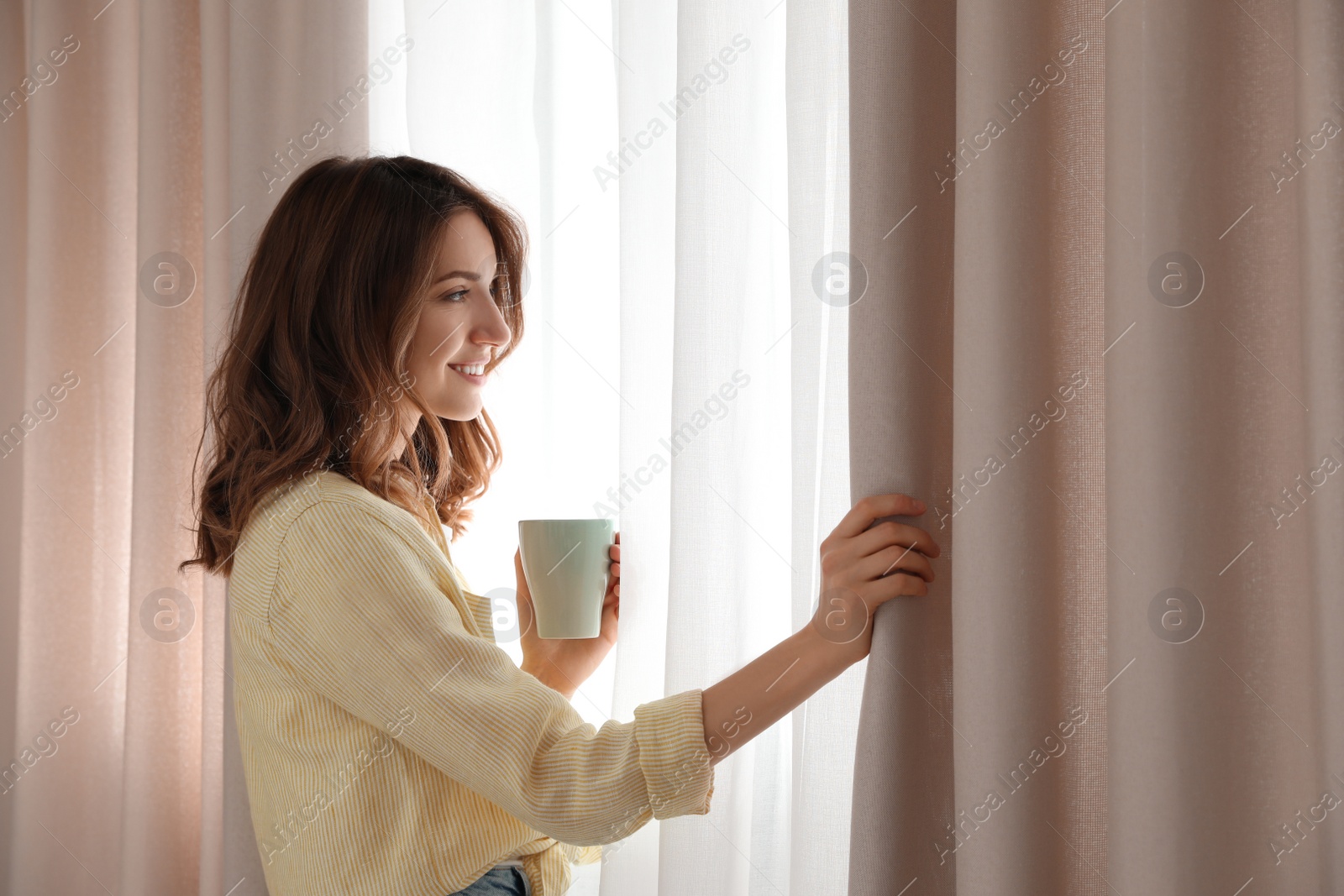Photo of Woman holding cup near window with beautiful curtains at home