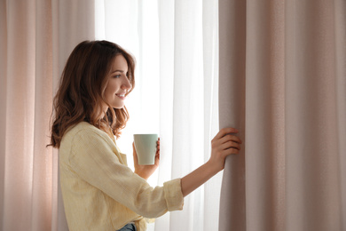 Woman holding cup near window with beautiful curtains at home