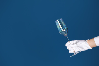 Person in white glove checking cleanliness of glass on blue background, closeup. Space for text