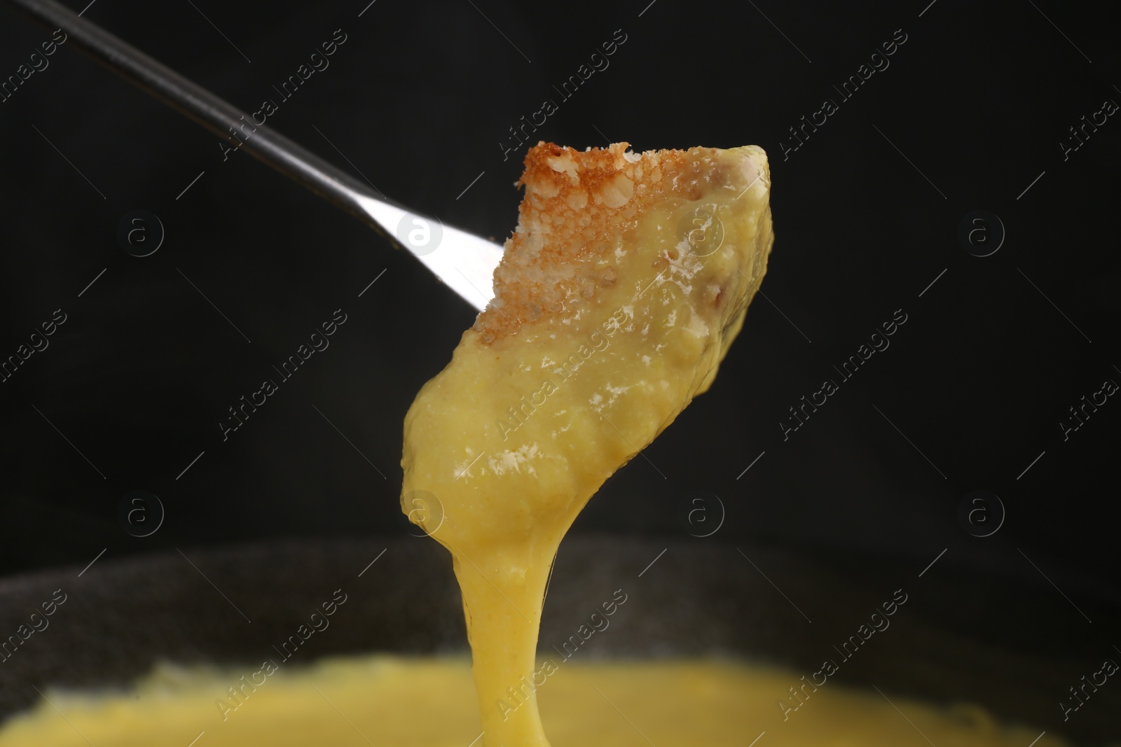 Photo of Dipping piece of bread into fondue pot with tasty melted cheese against dark gray background, closeup