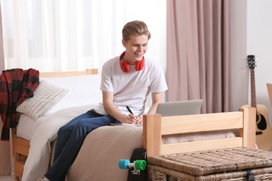 Online learning. Smiling teenage boy writing in notebook near laptop at home