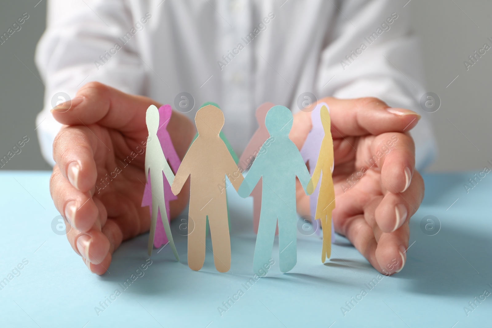 Photo of Woman protecting paper human figures on light blue table against grey background, closeup. Diversity and inclusion concept