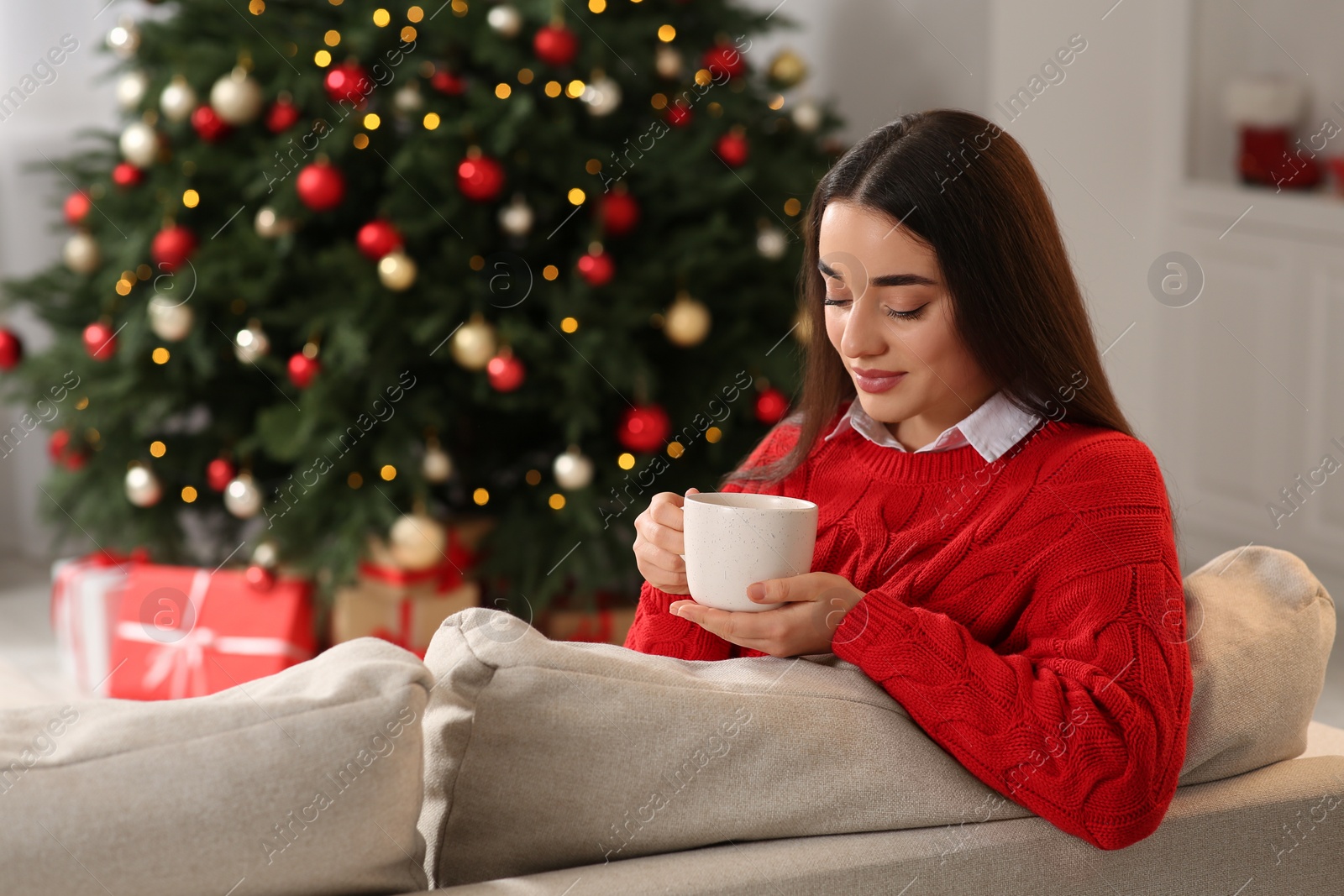 Photo of Woman holding cup of hot drink on sofa near Christmas tree indoors. Space for text