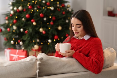 Photo of Woman holding cup of hot drink on sofa near Christmas tree indoors. Space for text