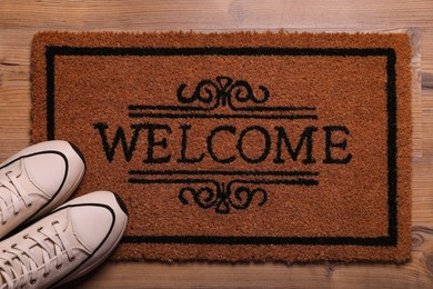 Photo of Door mat with word Welcome and stylish sneakers on wooden floor, flat lay