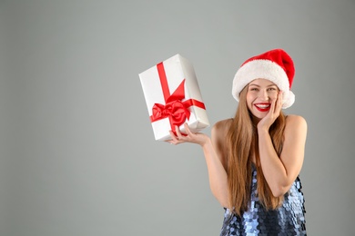 Young beautiful woman in Santa hat with gift box on grey background. Christmas celebration