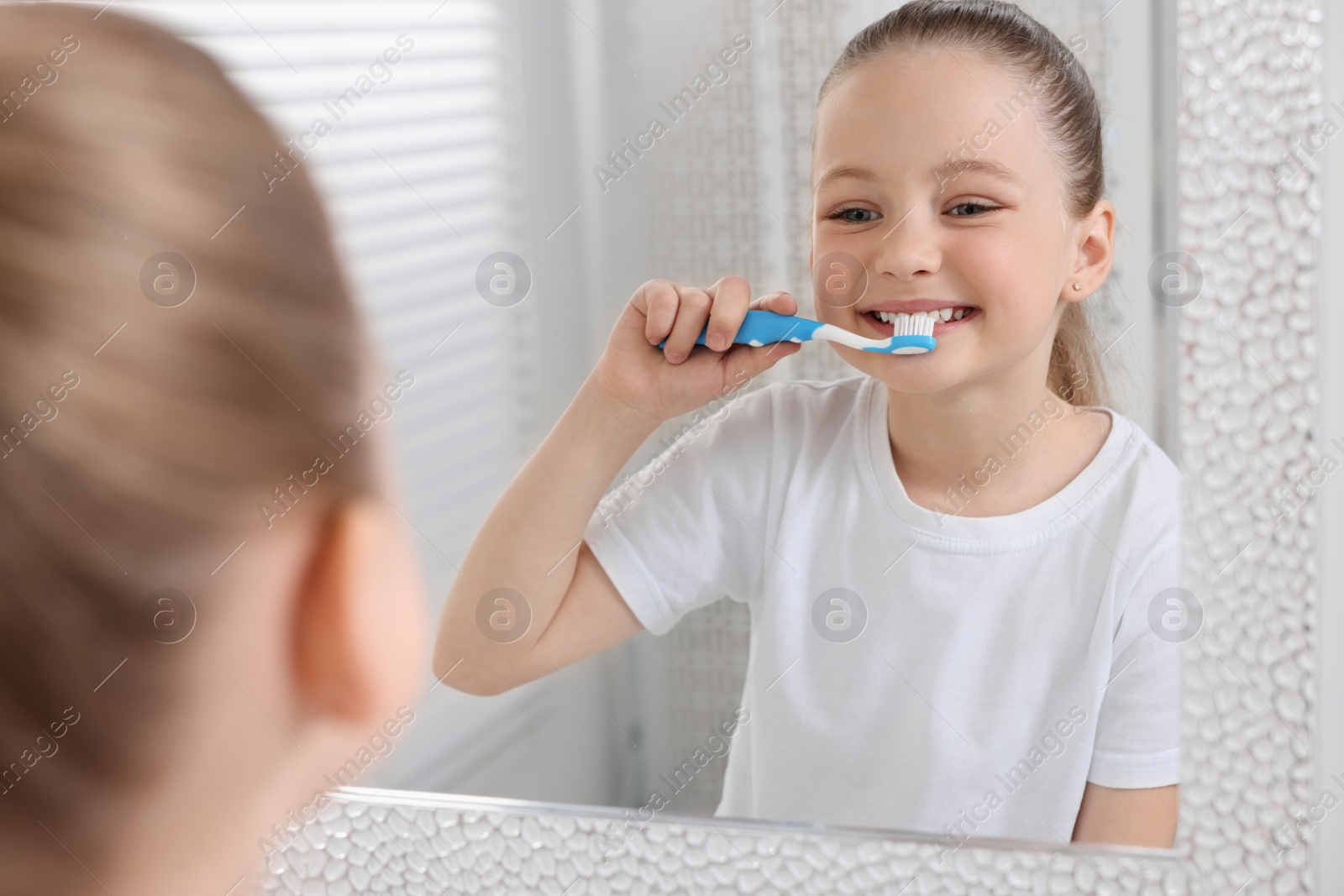 Photo of Cute little girl brushing her teeth with plastic toothbrush near mirror indoors