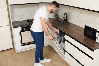 Handsome man opening dishwasher's door in kitchen