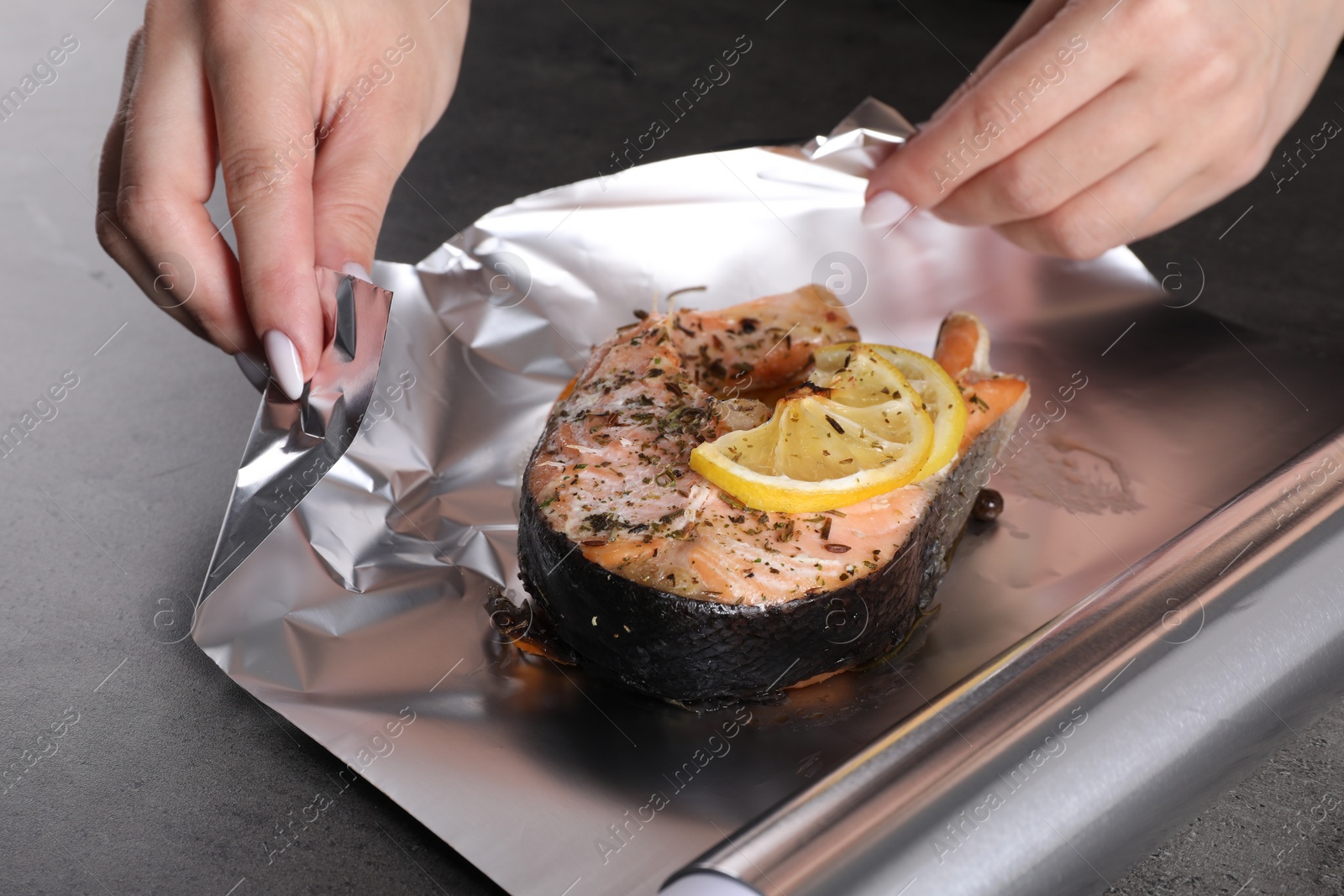 Photo of Woman wrapping tasty salmon with lemon and spices in aluminum foil at grey textured table, closeup