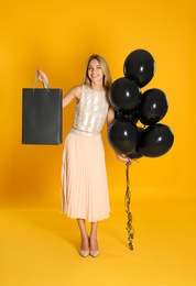 Happy young woman with balloons and shopping bag on yellow background. Black Friday Sale