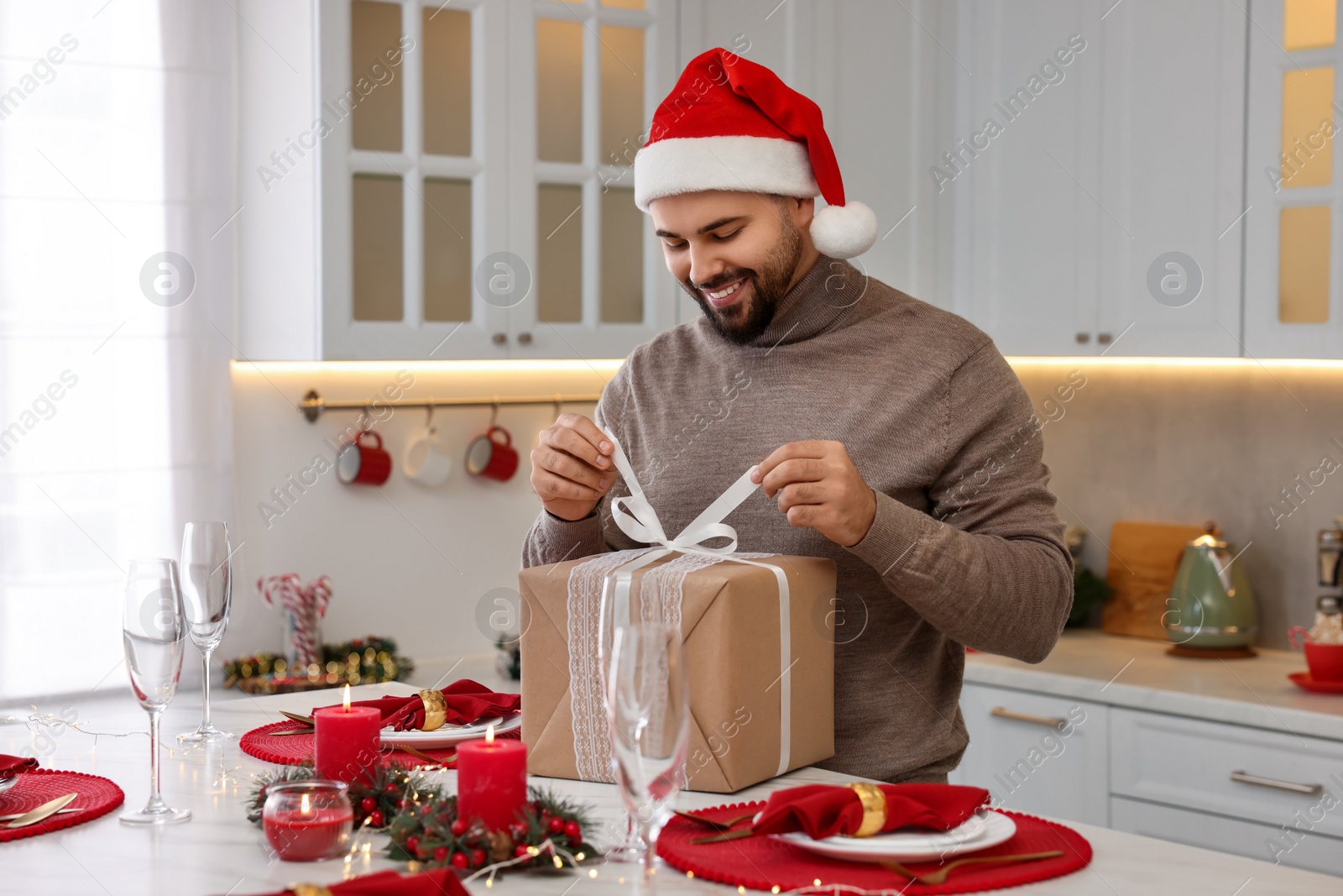 Photo of Happy young man in Santa hat opening Christmas gift at table in kitchen