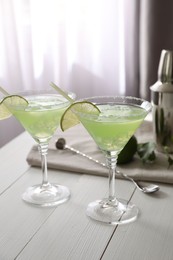 Delicious Margarita cocktail in glasses, lime and bar spoon on white wooden table