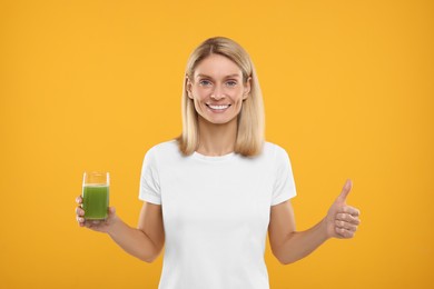 Photo of Happy woman with glass of fresh celery juice showing thumb up against orange background