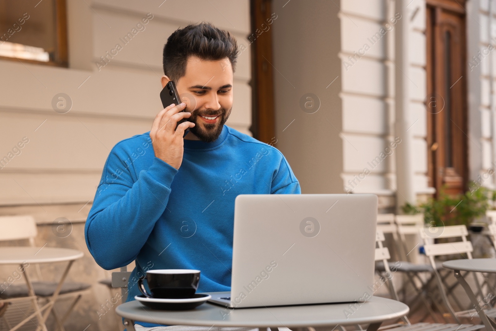 Photo of Handsome young man talking on smartphone while using laptop at table in outdoor cafe