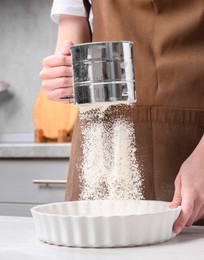Photo of Woman sieving flour into baking dish at table in kitchen, closeup