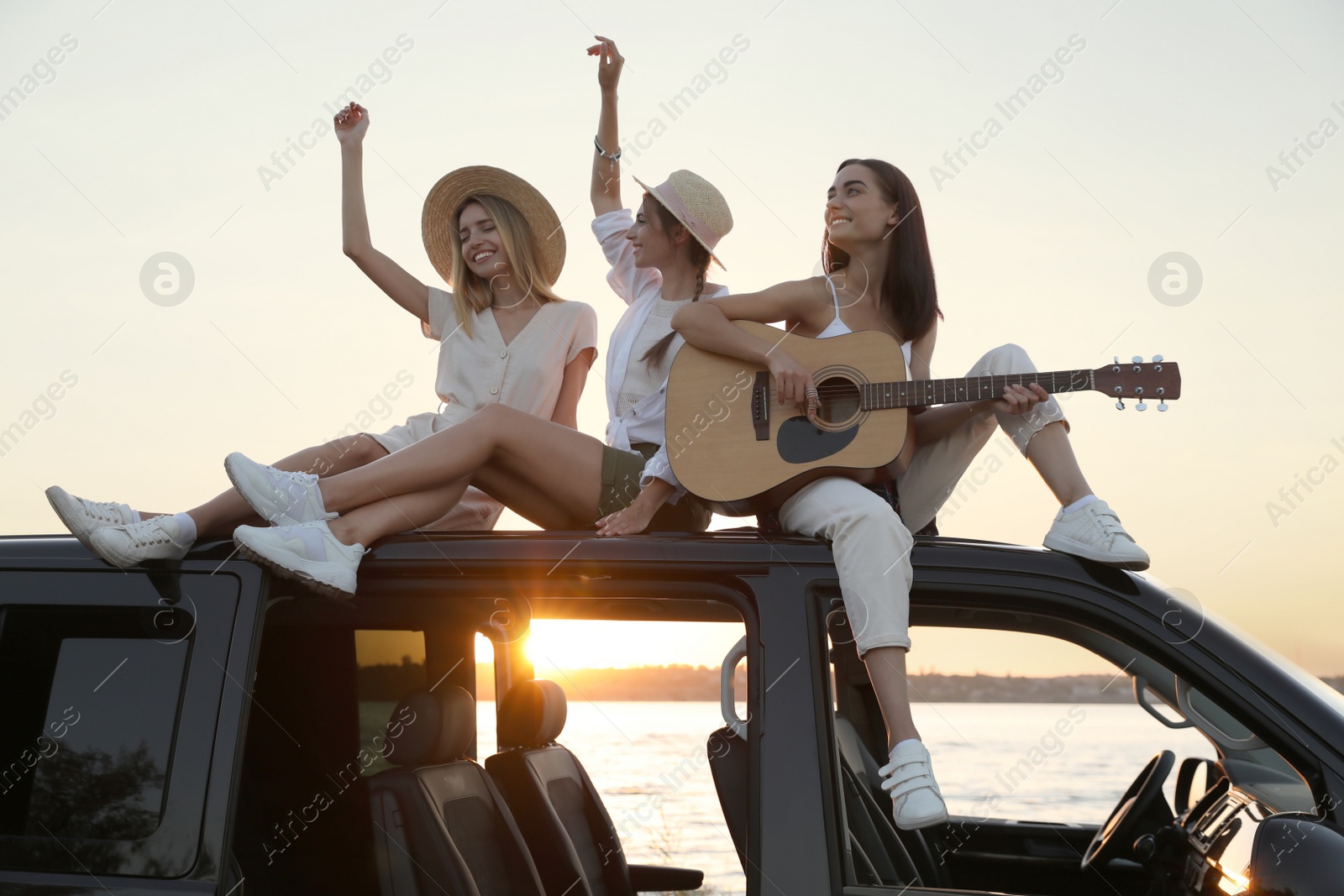 Photo of Happy friends with guitar sitting on car roof outdoors at sunset. Summer trip
