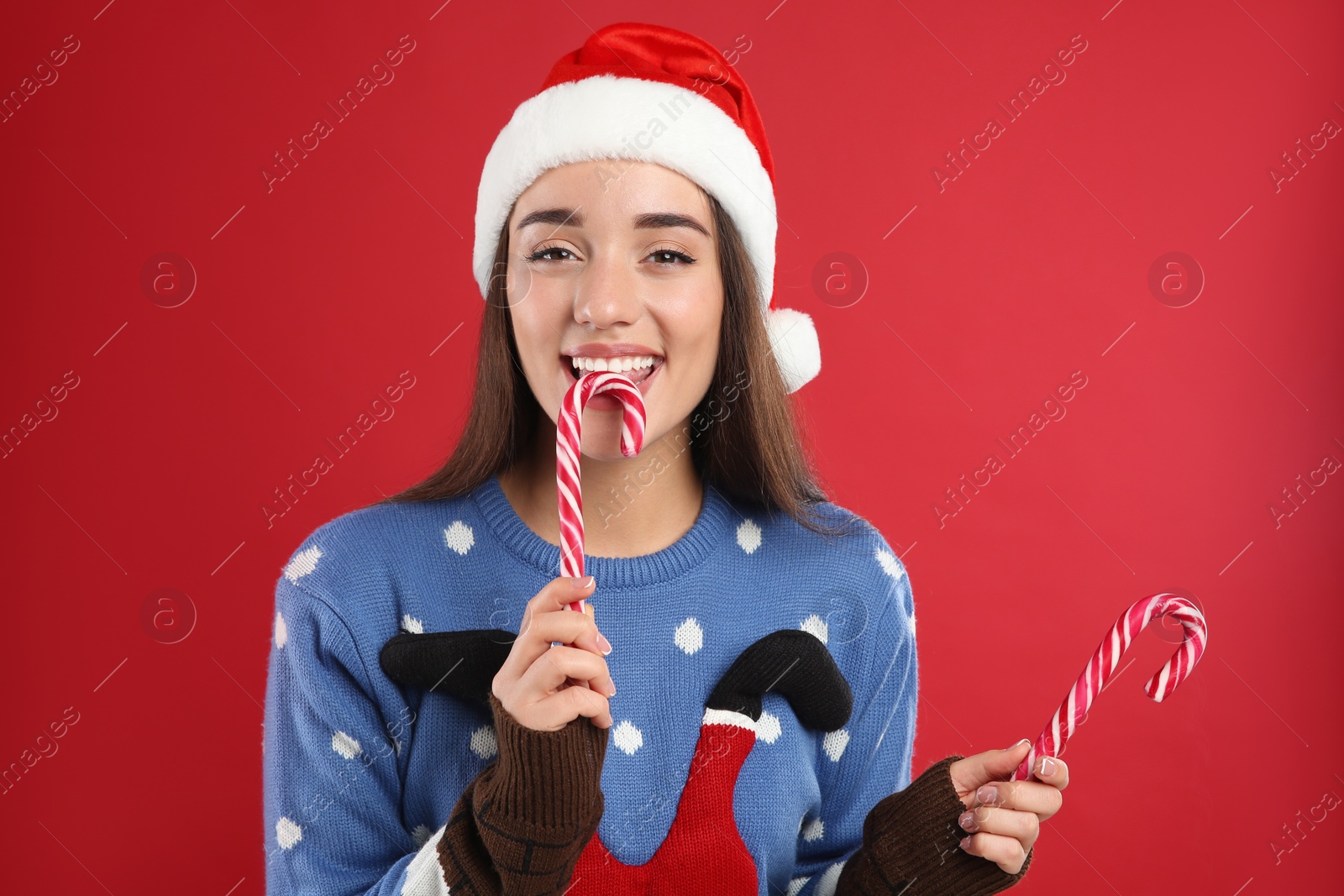Photo of Young woman in Christmas sweater and Santa hat holding candy canes on red background