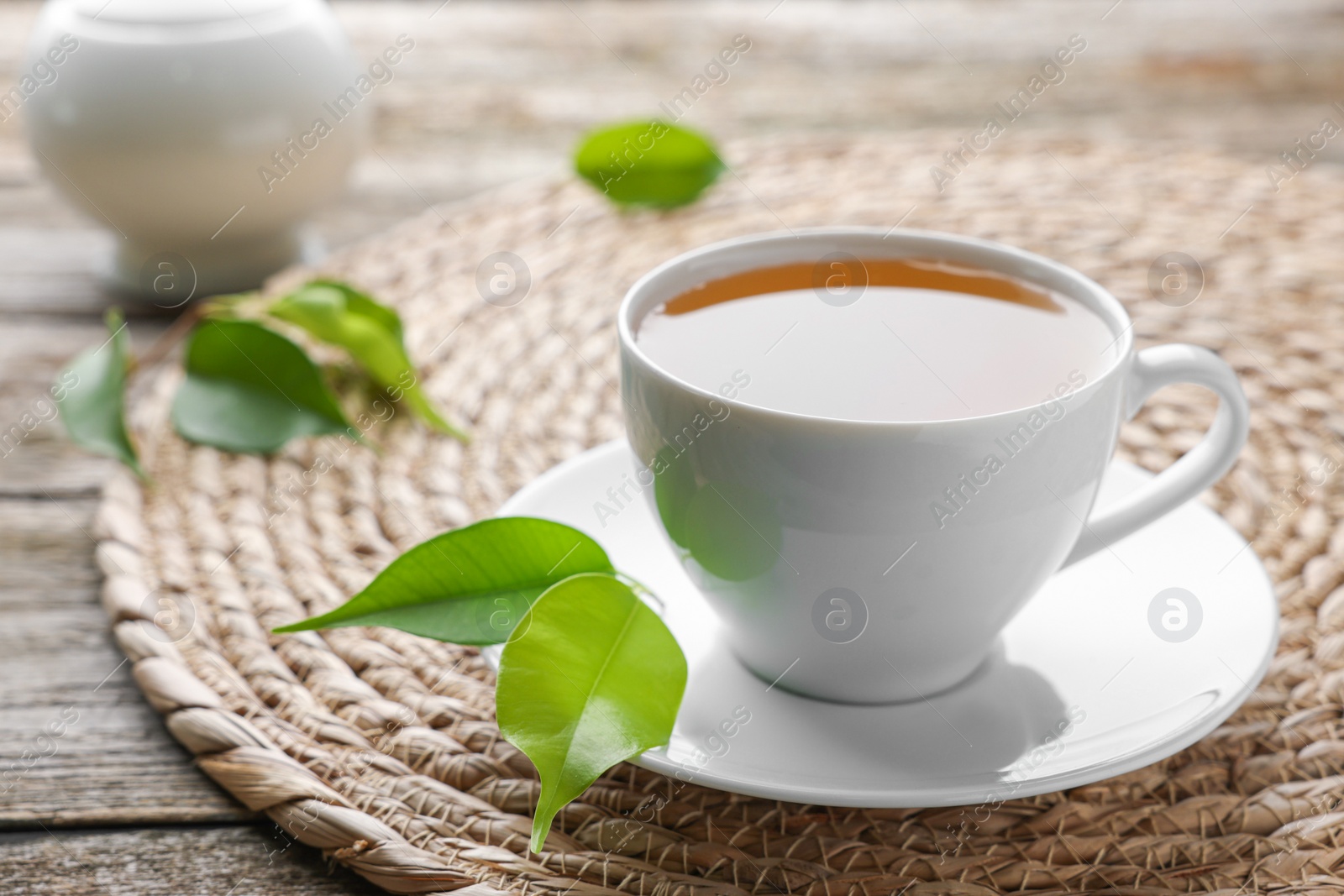 Photo of Green tea in white cup with leaves and wicker mat on table, closeup