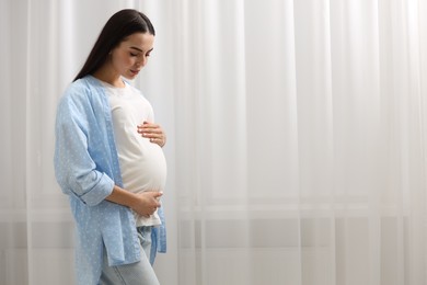 Pregnant young woman near window indoors, space for text