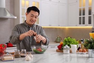 Photo of Cooking process. Man adding salt into bowl of salad at countertop in kitchen