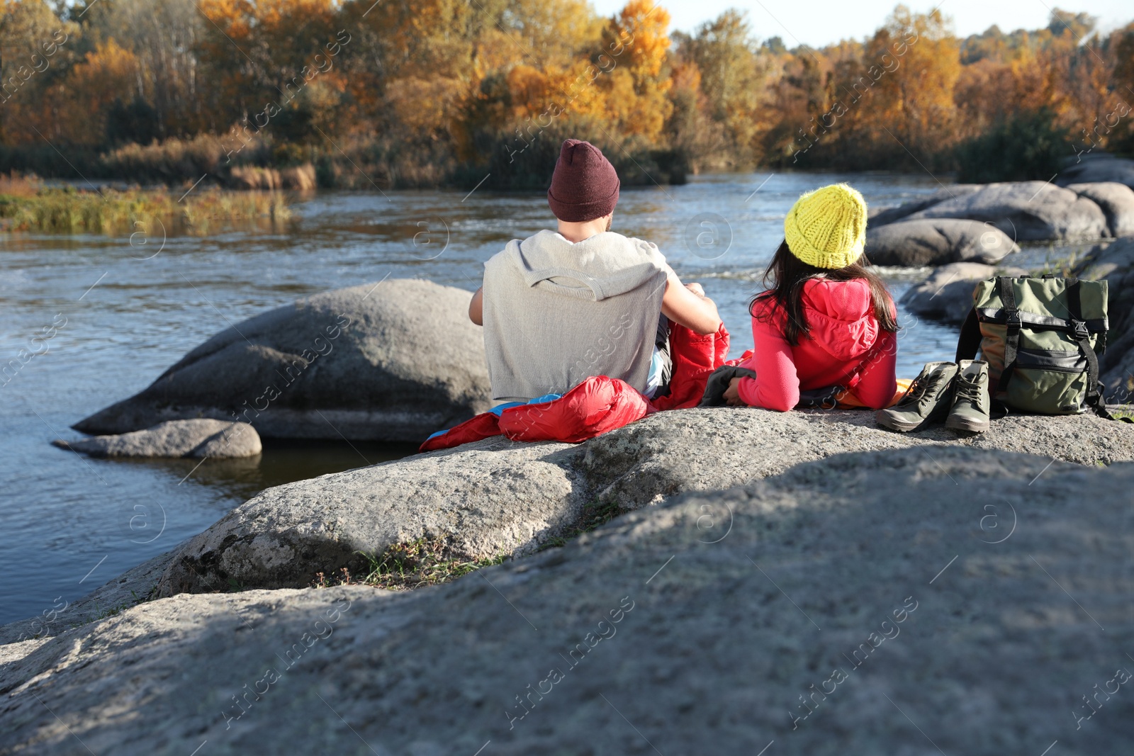 Photo of Couple of campers in sleeping bags sitting on rock near pond. Space for text