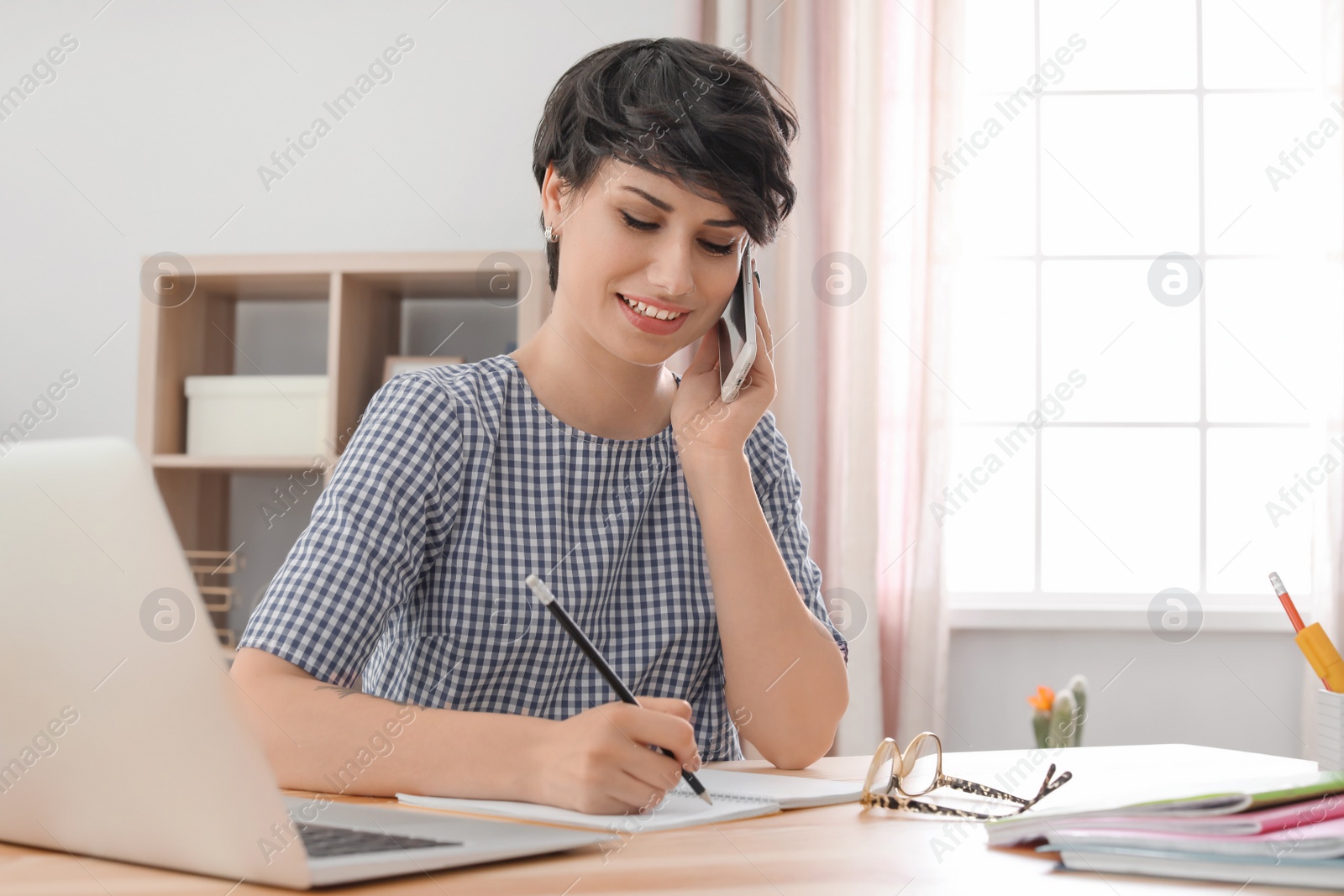 Photo of Young woman talking on mobile phone while working at desk. Home office