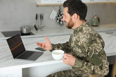 Photo of Happy soldier with cup of drink using video chat on laptop at white marble table in kitchen. Military service