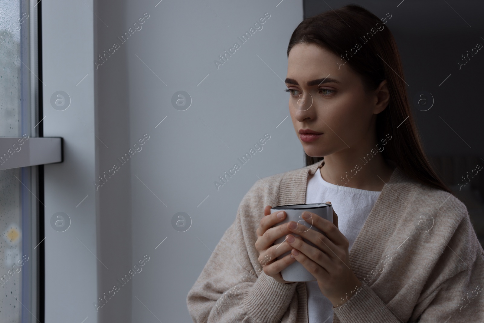 Photo of Melancholic young woman with drink looking out of window on rainy day, space for text. Loneliness concept