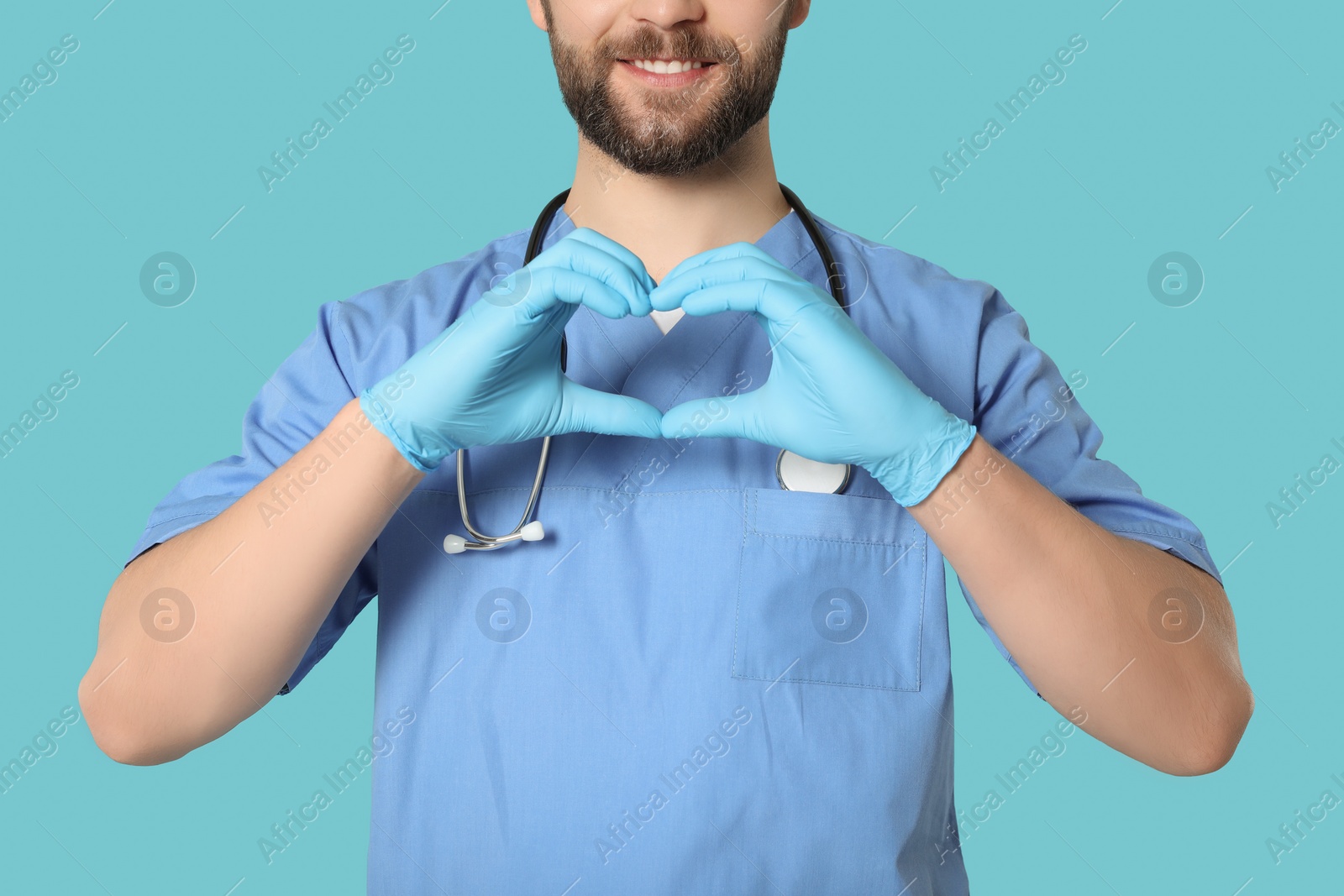 Photo of Doctor or medical assistant (male nurse) in uniform making heart with hands on turquoise background, closeup