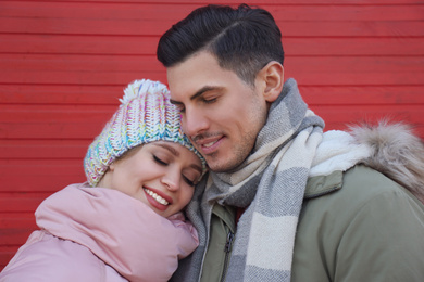Photo of Happy couple in warm clothes near red wooden wall outdoors. Christmas season