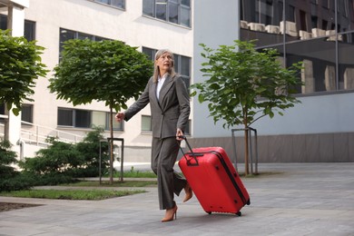 Photo of Being late. Worried senior businesswoman with red suitcase running outdoors