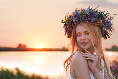 Photo of Young woman wearing wreath made of beautiful flowers outdoors at sunset