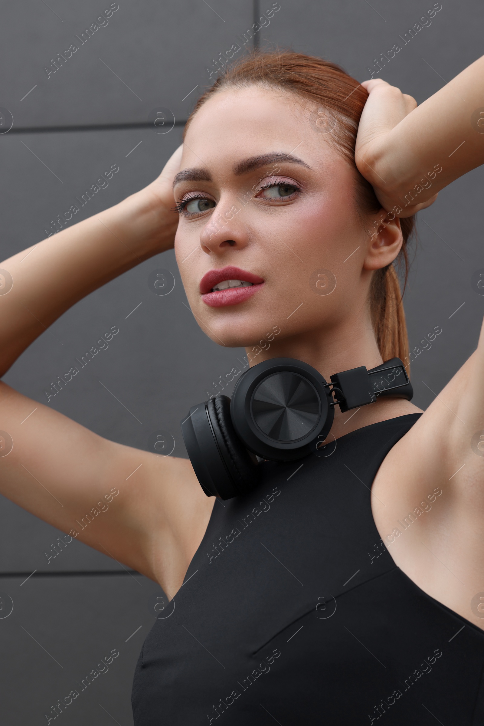 Photo of Beautiful woman in stylish gym clothes with headphones posing near dark grey wall on street, closeup