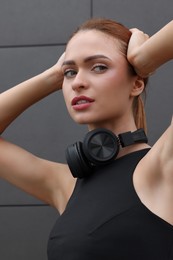 Beautiful woman in stylish gym clothes with headphones posing near dark grey wall on street, closeup