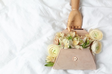 Photo of Stylish clutch with spring flowers and wooden mannequin hand on crumpled sheet, flat lay. Space for text