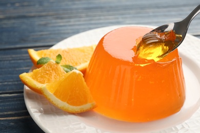 Spoon of tasty orange jelly over plate with dessert on blue wooden table, closeup